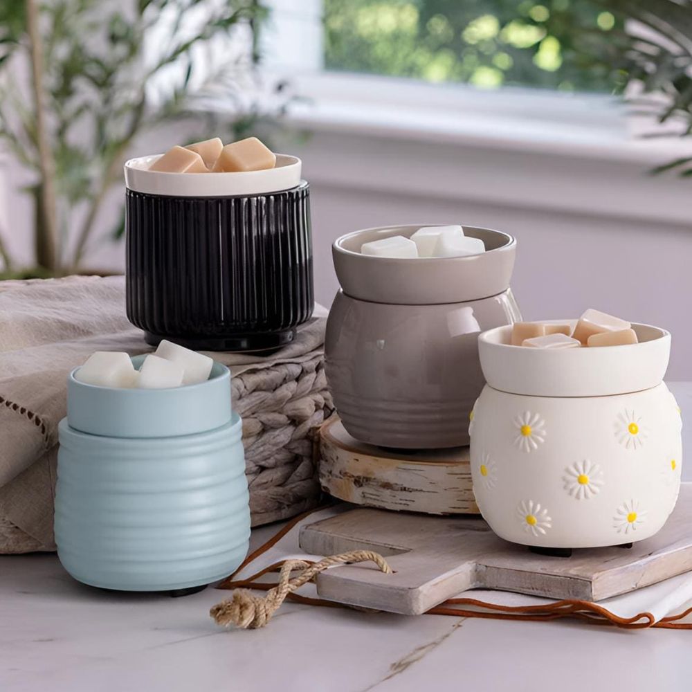Présentation de la collection de lampes électriques à plaque chauffante Candle Warmers avec les modèles RUCHE turquoise, Noir/Blanc strié, ORB gris et Marguerite, tous garnis de fondants de cire parfumés sur une table décorée.