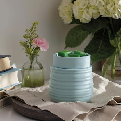 Vue d'ambiance de la lampe parfumée avec plaque chauffante RUCHE turquoise en céramique texturée, remplie de cire verte et posée sur un linge en lin sur une table en bois.